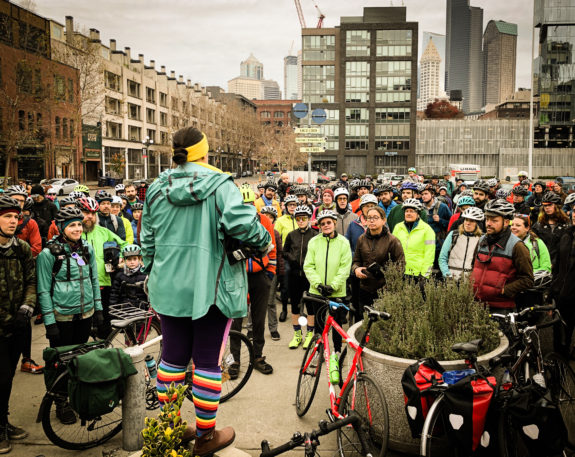 Photo of woman standing outside addressing a large crowd of people with bikes. 