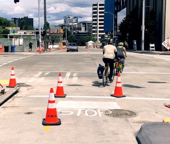 Two people ride bikes across an intersection, but the bike lane they were in does not continue on the other side. A car faces them.