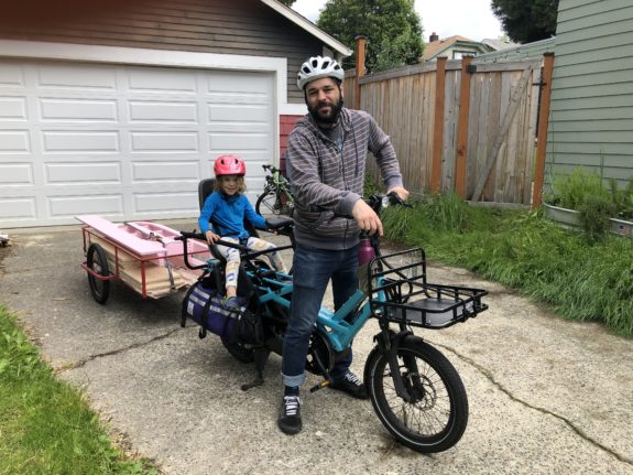 Photo of a person on a bike with a kid in a child seat and a trailer full of lumber.