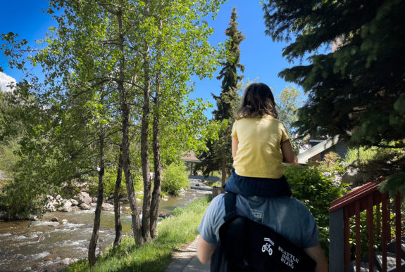A kid on an adult's shoulders walking next to a creek. 