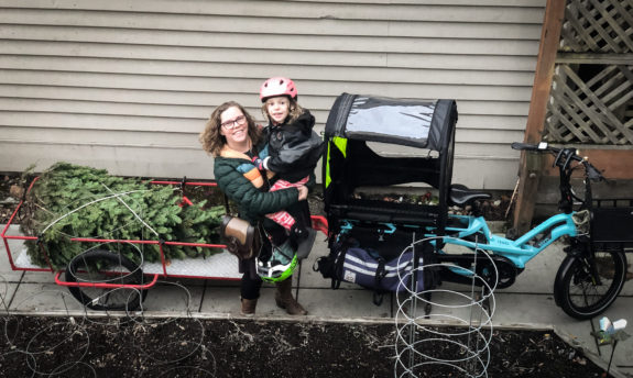 A mom and kid posing in front of a family cargo bike with a christmas tree in a trailer.