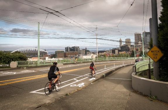People biking in bike lanes on the Jose Rizal Bridge with downtown Seattle in the background.