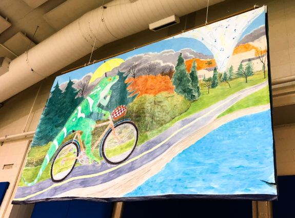 A mural hanging from the ceiling depicting a lizard riding a bicycle on the green lake path while holding an umbrella.
