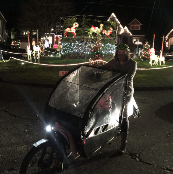 A person and kid on a cargo bike in front of some holiday lights.