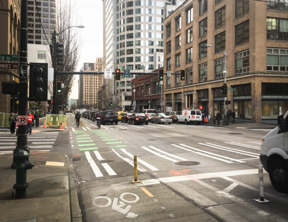 A car waits to turn at an intersection with people biking in a protected bike lane and walking in a crosswalk. There is a no turn on red sign.