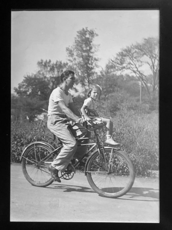 Old black and white photo of a child sitting on a seat on the handlebars while a man bikes. 