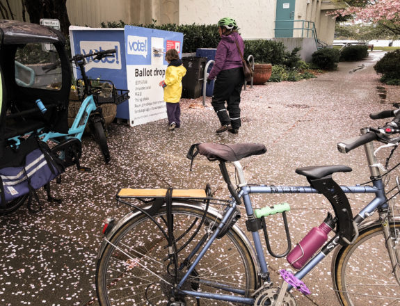 Photo of a road bike with a top tube child seat and an electric cargo bike. A child and adult are in the background putting ballots into a ballot box.