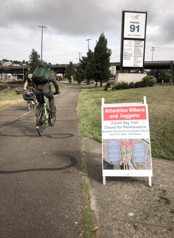 Two people bike past a sign warning of the upcoming detour.