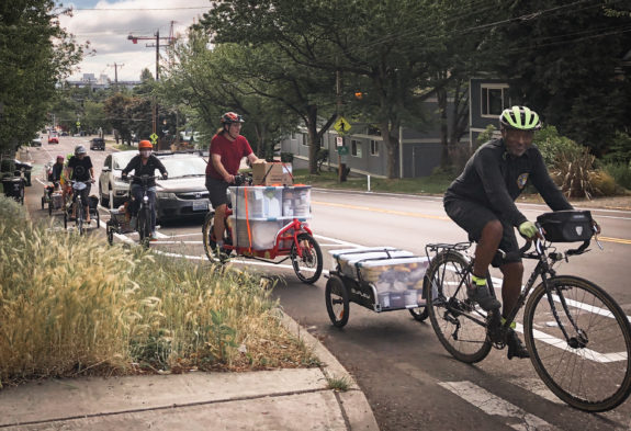 A half dozen people biking up a hill with boxes of food on trailers or cargo bikes.
