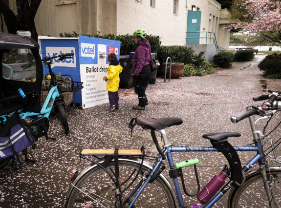An adult and a kid putting a ballot in a ballot box with bikes in the foreground.