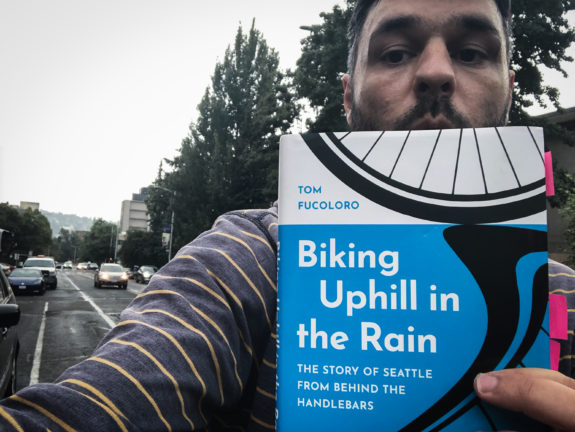 Selfie of the author in a rainy bike lane holding a copy of his book Biking Uphill in the Rain.