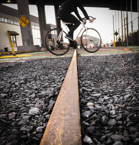 Photo from near the ground looking down a railroad track as someone bikes across. The Ballard Bridge is overhead.