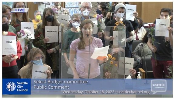 Screenshot from the Seattle Channel telecast of a budget hearing with many people standing behind a speaker holding signs with people's names.