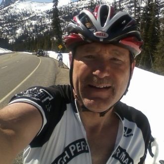 Selfie of a man in a helmet and cycling jersey on the side of a road surrounded by snow.