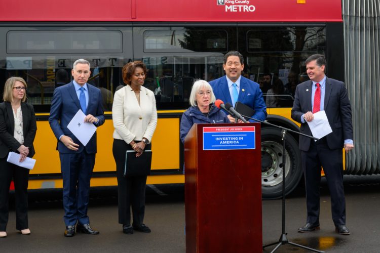 Six people standing in front of a bus while Patty Murray speaks at a podium with the words President Joe Biden Investing in America.