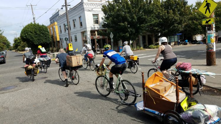 Photo of a group of people biking while carrying a variety of items, including one person with a desk in a bike trailer.